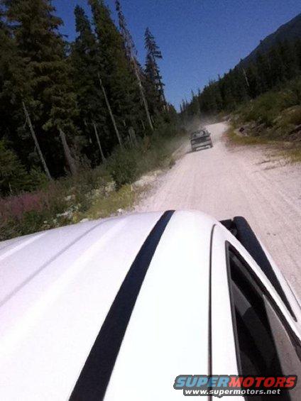 View from inside a white truck looking down a dusty dirt road with another truck ahead. Trees line the road