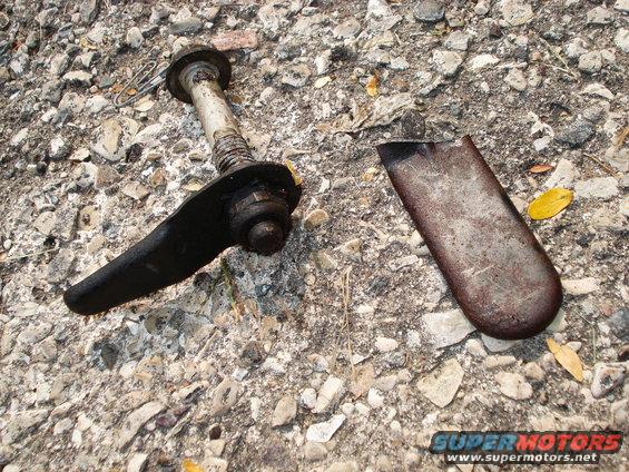 Broken black control arm with bolt and washer, next to a rusted metal plate on gravel