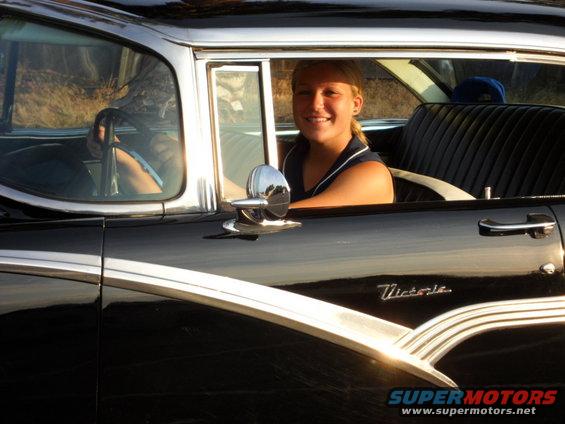 Black 1956 Ford Fairlane Victoria side view with chrome trim and a woman driving