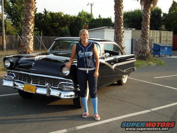 Woman in baseball jersey and pants stands by a black 1956 Ford Fairlane with a yellow license plate
