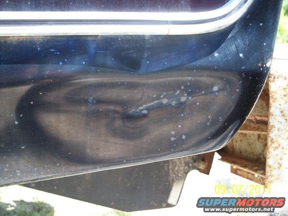 Dark blue vehicle body panel with a dent and white specks, showing a chrome trim piece above. A rusty metal frame is visible on the right