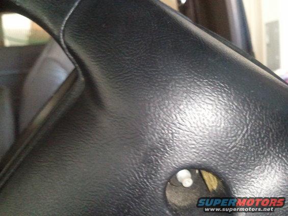 Close-up of a black steering column cover with a hole showing a white plastic piece. Supermotors.net logo visible