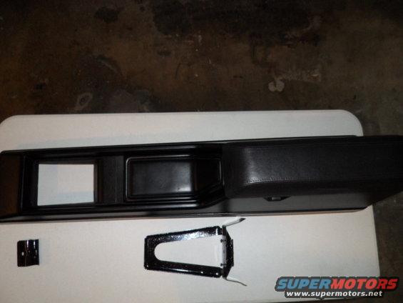 Black car interior console with two rectangular openings and a padded armrest, next to a metal bracket