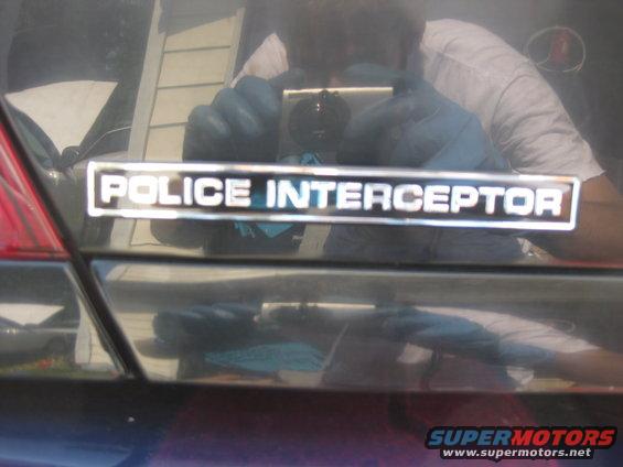 Chrome 'Police Interceptor' badge on a dark car trunk, reflecting a person holding a camera