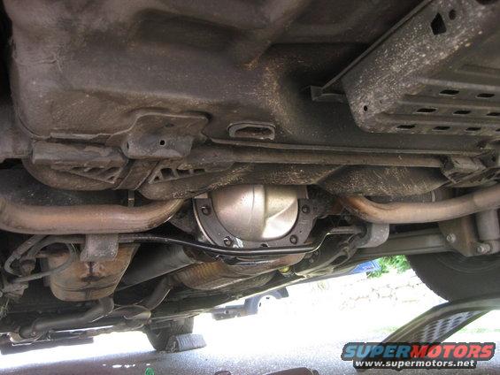 Underside view of a car's rear axle and exhaust system. There's a SuperMotors.net watermark