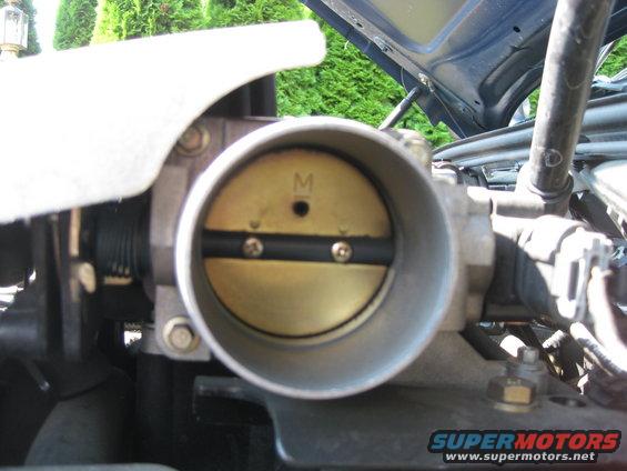 Close-up of a car's throttle body with the butterfly valve visible. There's an 'M' marking on the valve
