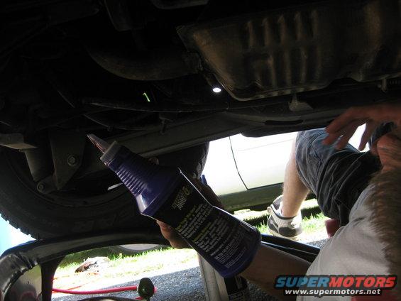 Person performing an oil change under a car, holding a bottle of oil additive