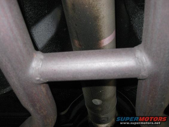 Underside view of a vehicle's exhaust system with a crossbar and drive shaft visible. The metal parts show some wear and dirt