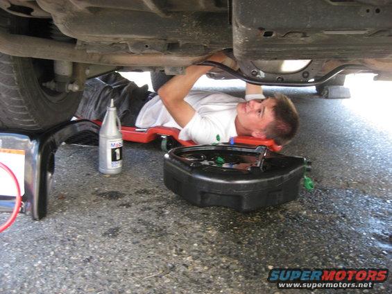 Person on creeper under vehicle, with oil drain pan and Mobil 1 oil bottle