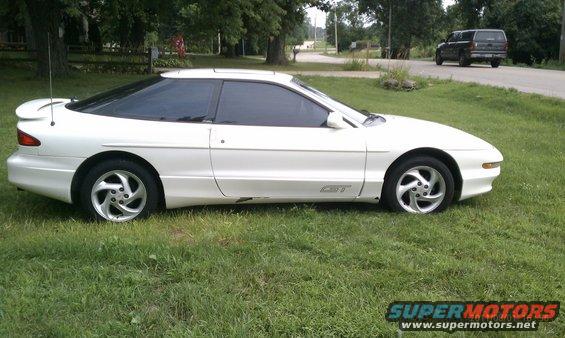 White 1994 Ford Probe GT parked on grass. A black SUV is visible on the road in the background