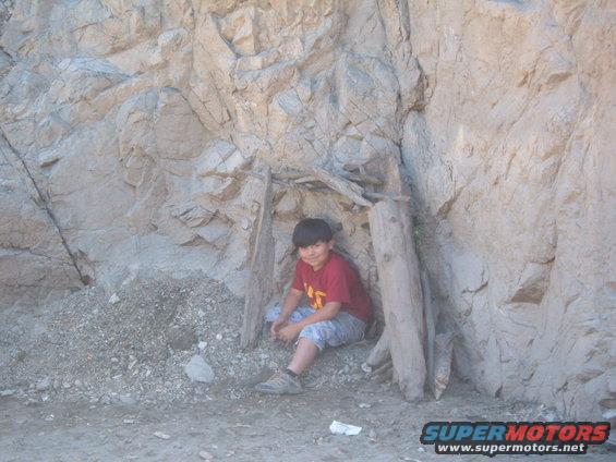 Kid sitting in a makeshift shelter against a rock face. There's a pile of rubble to the left and some wooden posts supporting the roof