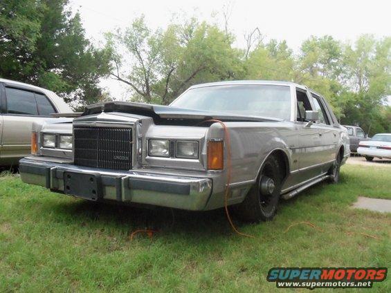 Silver Lincoln Town Car with hood open, missing front passenger wheel, parked on grass