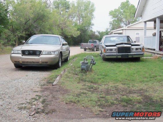 Gold sedan with quad headlights parked on gravel, silver sedan with hood up on grass, pickup truck in background