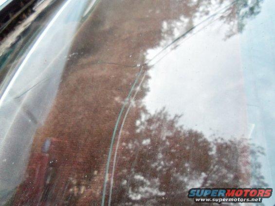 Cracked windshield with reflection of trees and brick building. Supermotors.net logo visible