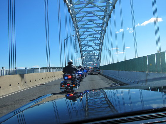 Police motorcycles and a car driving on a bridge under a clear blue sky