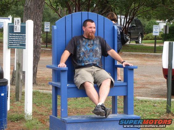 Man in a giant blue Adirondack chair with a dark SUV in the background