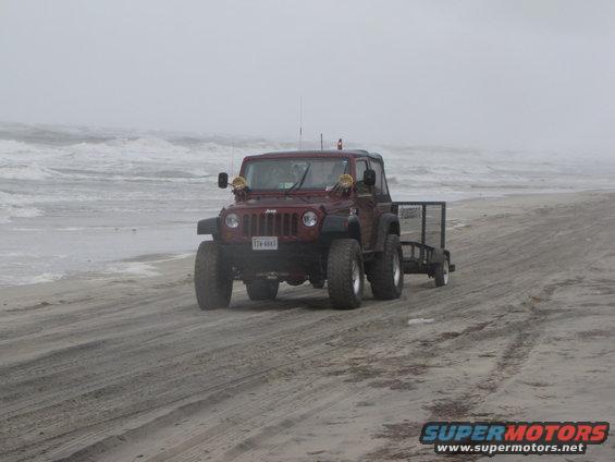 Red Jeep Wrangler with a trailer driving on a beach with ocean waves in the background