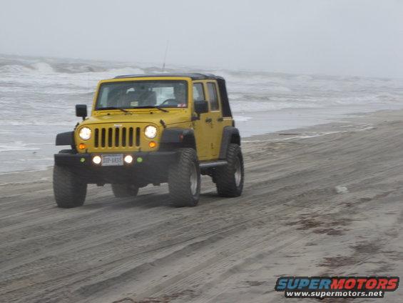 Yellow Jeep Wrangler driving on a sandy beach with waves in the background