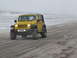 1979 Ford Bronco - A yellow Jeep Wrangler drives along a sandy beach on an overcast day.