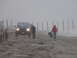 1979 Ford Bronco - A blue SUV drives on a sandy beach during foggy weather, with two individuals walking ahead.