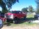 1989 Ford Bronco - A red pickup truck with a lifted suspension and large tires is parked on a grassy area next to other vehicles.