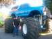 1989 Ford Bronco - A man stands beside a blue monster truck with 'High Horse' painted on the side, oversized tires and suspension.
