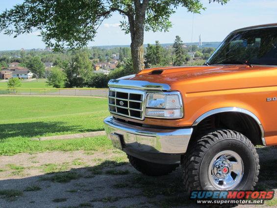 Front corner of orange pickup truck with chrome grille and bumper. Hood scoop visible