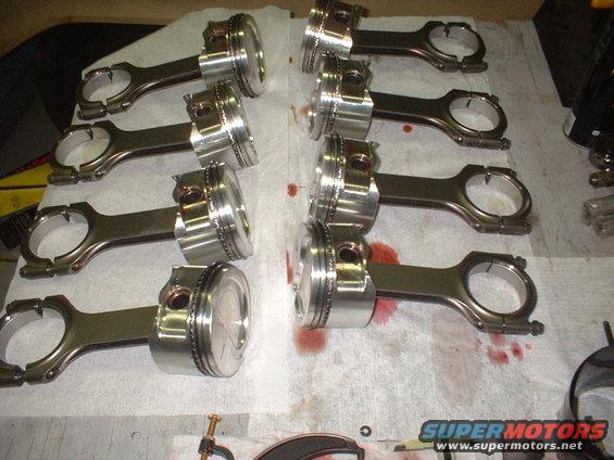 Eight pistons and connecting rods laid out on a surface with oil stains