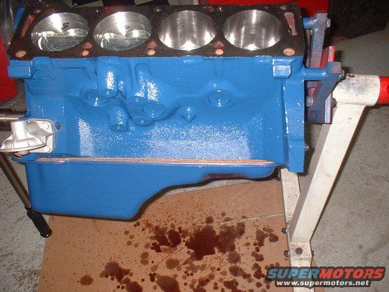 Blue engine block with four visible pistons, mounted on an engine stand. Oil stains on cardboard below