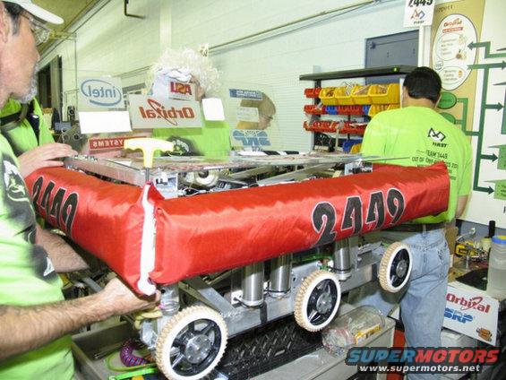 Robot with red bumper labeled 2449, four wheels, and metal frame, held by people
