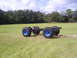 1992 Ford Bronco - This vehicle chassis, equipped with oversized tires and striking blue rims, sits on a wide expanse of grass under a partly cloudy sky.