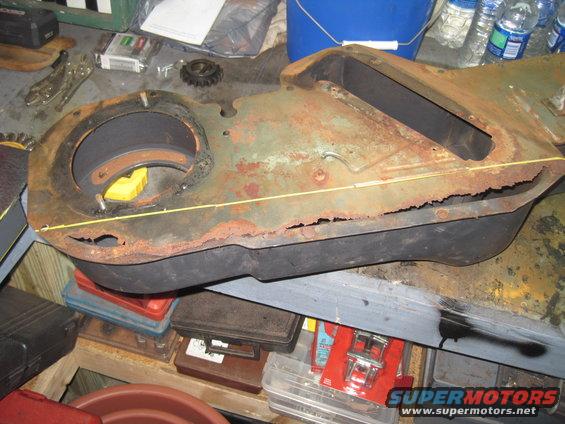 Heavily rusted metal component with circular opening and rectangular cutout, resting on a workbench with tools and supplies