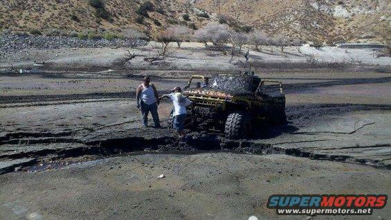 Yellow off-road vehicle covered in mud, stuck in a muddy ditch with two people standing nearby