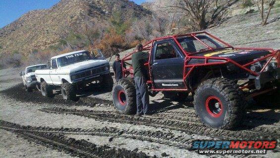 Two off-road trucks, one white pickup with 'THE FEW' on windshield, and a black buggy with red roll cage, on a dirt track