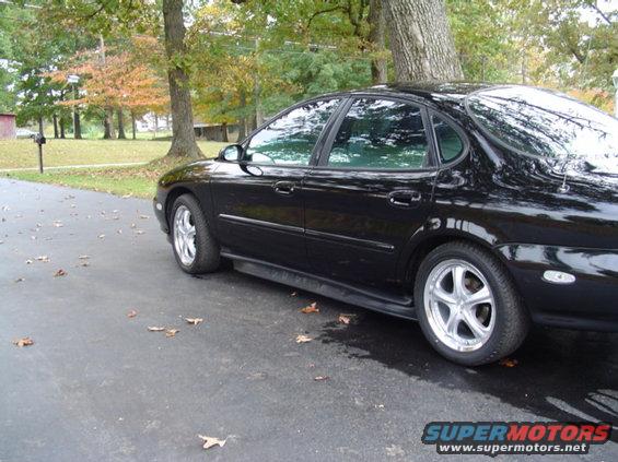 Black sedan parked on asphalt driveway with trees and fallen leaves in the background. It has aftermarket wheels