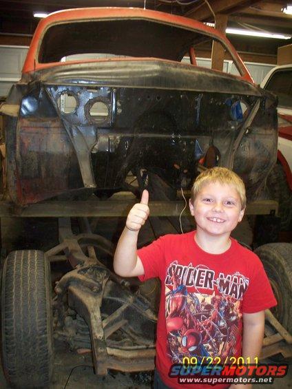 Kid gives thumbs up in front of a disassembled car body, possibly a Ford Bronco, in a garage
