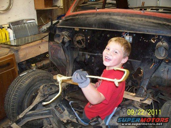 Child holding a metal rod inside a disassembled vehicle's engine bay, with a tire visible