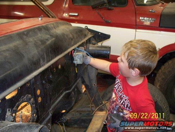 Boy in red shirt works on car engine bay; red Bronco visible in background