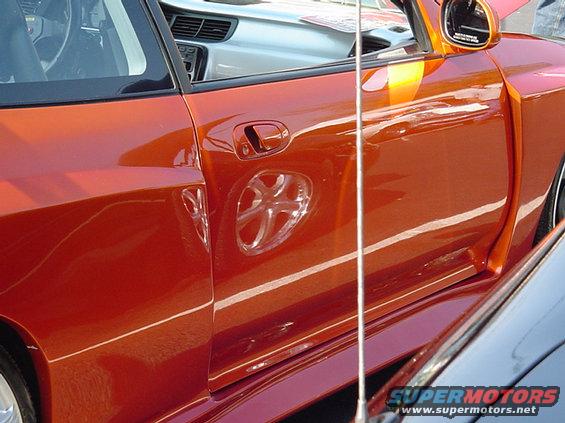Orange car door with a clear plastic accent in the wheel well area. A silver car is parked next to it