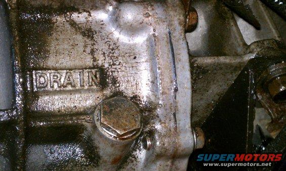 Close-up of a greasy transmission housing with the word DRAIN stamped into it and a hexagonal drain plug