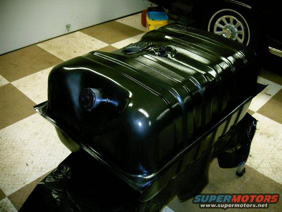 black fuel tank, looks freshly painted, sitting on black plastic sheeting. A car wheel is visible in the background