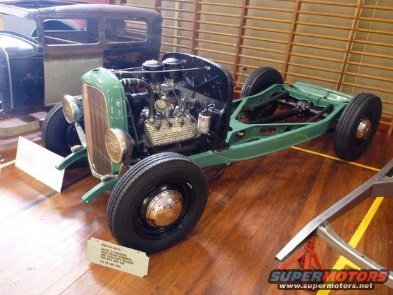 Green hot rod chassis with V8 engine and front grille. Another car body is behind it