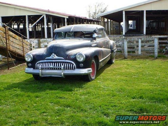 Old dark sedan with a prominent grille parked on grass in front of farm buildings