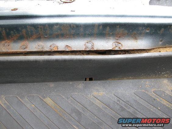 Close-up of rust on a dark green vehicle panel above a textured black plastic trim piece