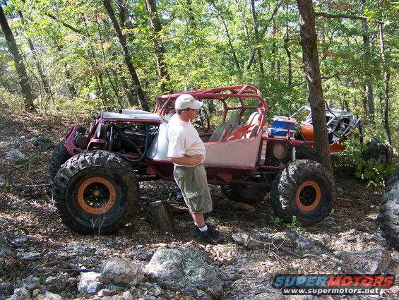 Man standing next to a custom red off-road buggy with large tires. Another buggy is visible behind it