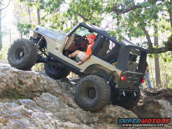 Jeep Wrangler with large tires climbing over rocks, driver wearing an orange hat
