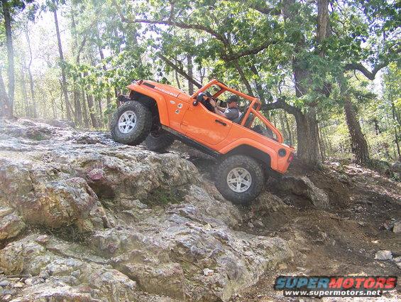 Orange Jeep Wrangler Rubicon climbing a rocky incline in a wooded area. "Rubicon" is visible on the hood