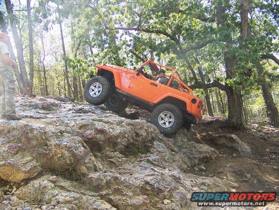 Orange Jeep Wrangler climbing a rocky hill with a person standing nearby