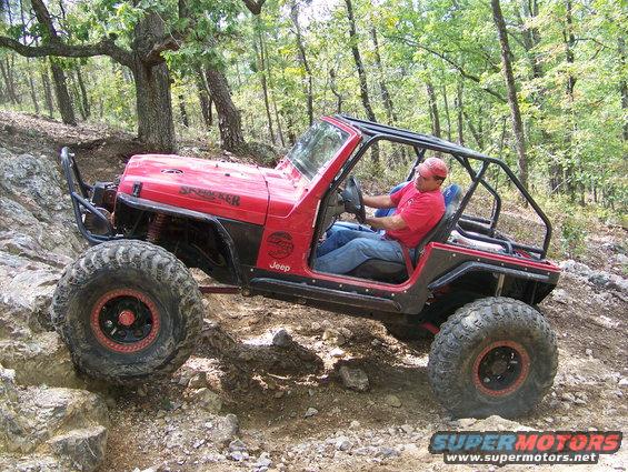 Red Jeep Wrangler with a roll cage driving over rocks on a wooded trail