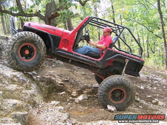 Red Jeep Wrangler with roll cage climbing over rocks, driver wearing a red cap and shirt